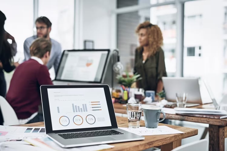 Closeup shot of a laptop with graphs on the screen and a team working in the background