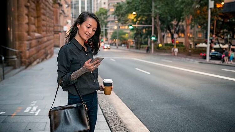 Telecommunications: A woman stands a sidewalk holding a coffee and looking at her mobile phone.