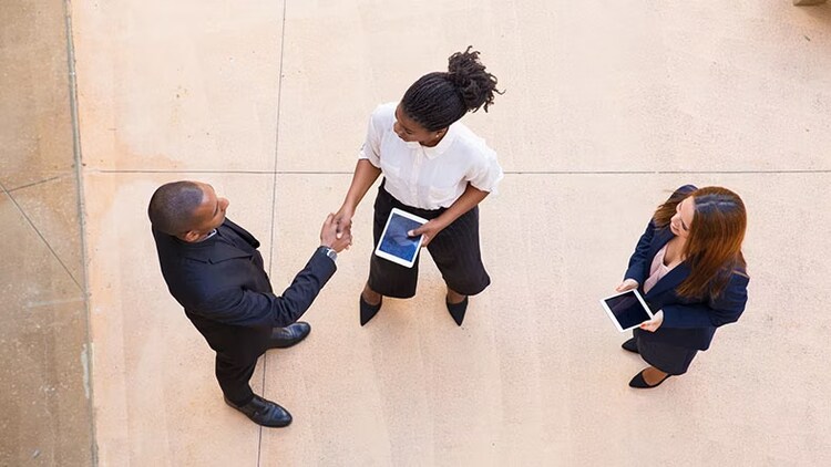 Employees holding tablets greet a customer with a handshake.