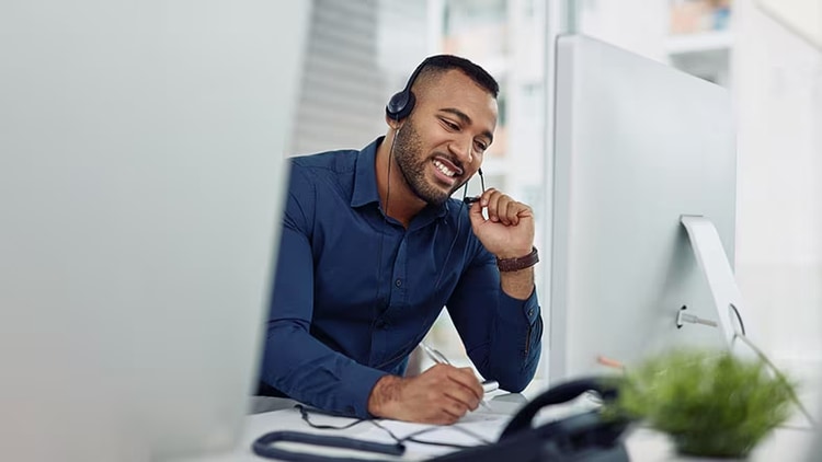 A customer service rep wearing a headset jots notes in front of a computer monitor.