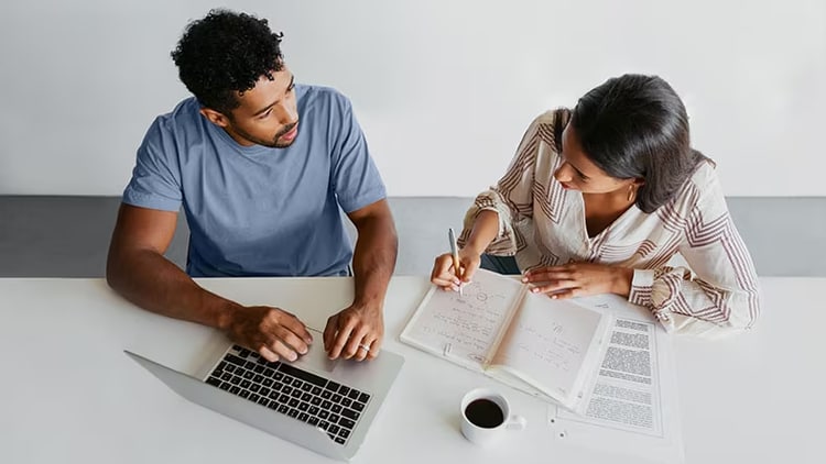 Two workers sit at a table with a laptop and a notebook.