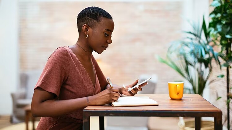 Supporting hybrid work: A woman looks at a phone while writing in a notebook.