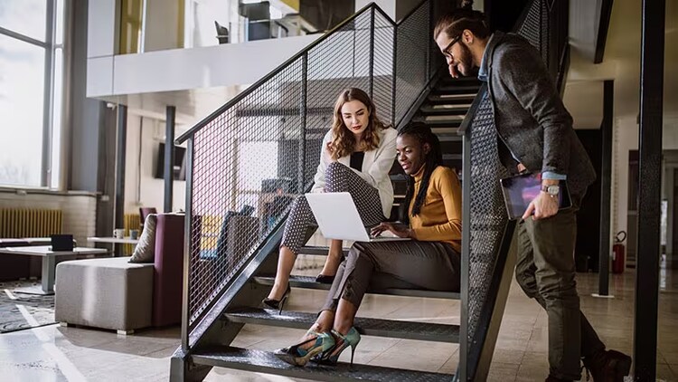 Privacy management: A man holding a tablet leans over a stair rail to look at a woman's laptop computer while another woman on the stairs looks on.