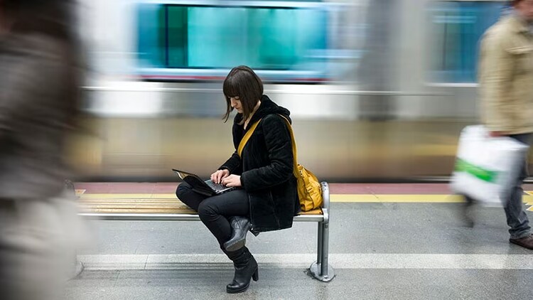 Managing risk and security: A woman sits on a bench using a laptop at a train station.