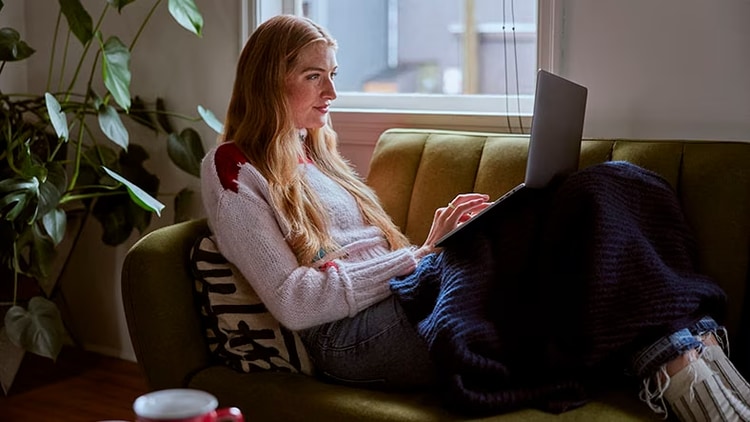Microsoft Teams collaboration: A woman sits on a couch working on a laptop.