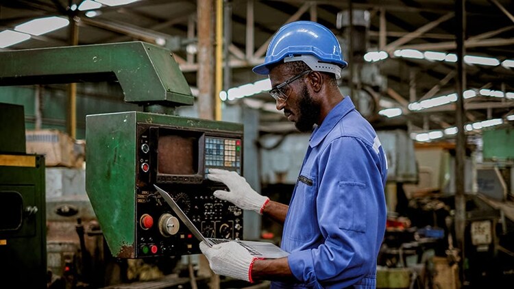 IT and operational technology asset management in manufacturing: man in factory looking at laptop while working machine