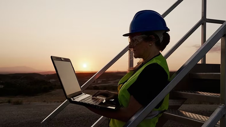 Field service: woman wearing hard hat looking at laptop outdoors at sunset