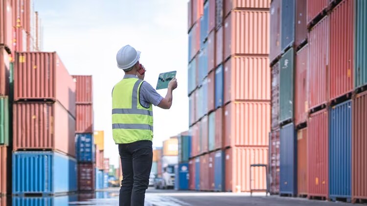 Finance and supply chain: man in hard hat among shipping containers talking into walkie-talkie