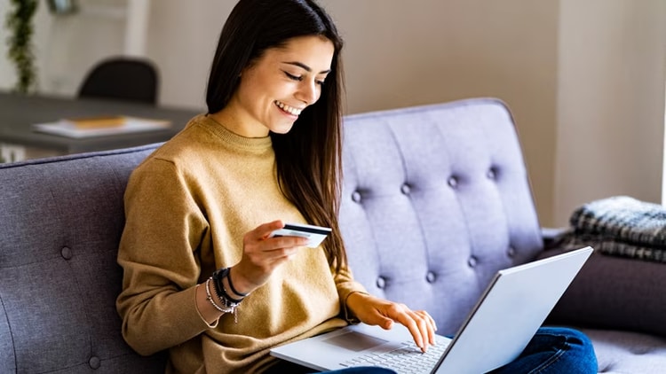 technology risk: smiling woman on couch holding a credit card, looking at a laptop