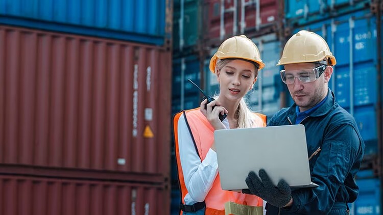 Supply chain resilience: 2 workers in hard hats looking at a laptop in front of shipping containers
