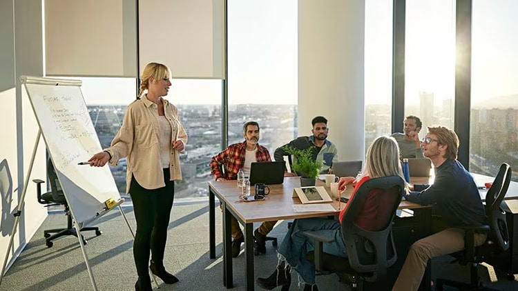 Technical project manager pointing at a whiteboard in front of five seated employees in a corner conference room