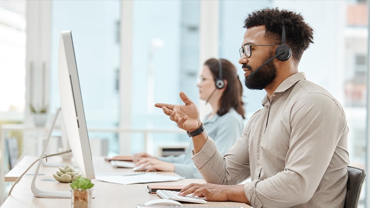 A support agent wearing a headset pointing to his computer with another support agent in the background
