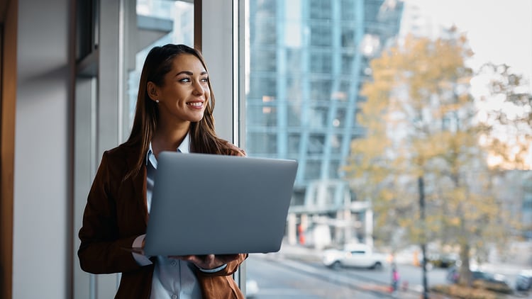 AI questions: smiling woman holding a laptop looking out an office window
