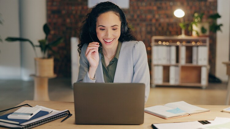Woman wearing a headset smiling at an open laptop on her desk