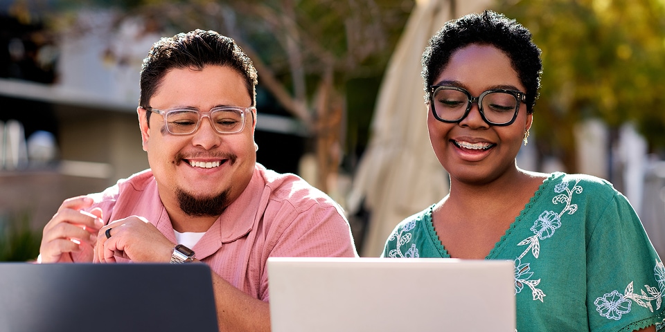 Deux apprenants sourient tout en travaillant ensemble sur un ordinateur portable à l’extérieur lors d’une séance des journées d’apprentissage IA ServiceNow.