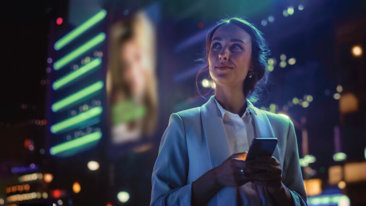 KI-Revolution: Frau, die nachts draußen ein Smartphone in der Hand hält und vor hohen, beleuchteten Gebäuden in die Ferne schaut