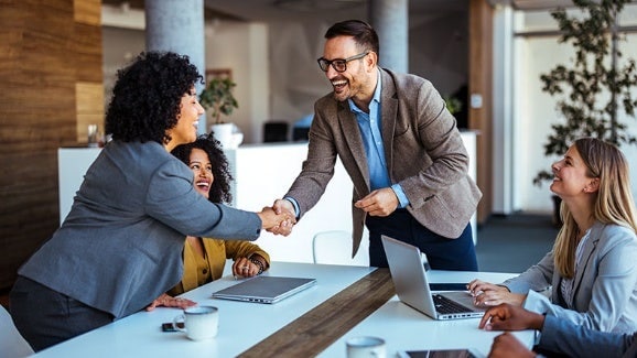 Vier Personen in einem modernen Büro; zwei stehen und geben sich die Hand, zwei sitzen am Tisch mit Laptops.