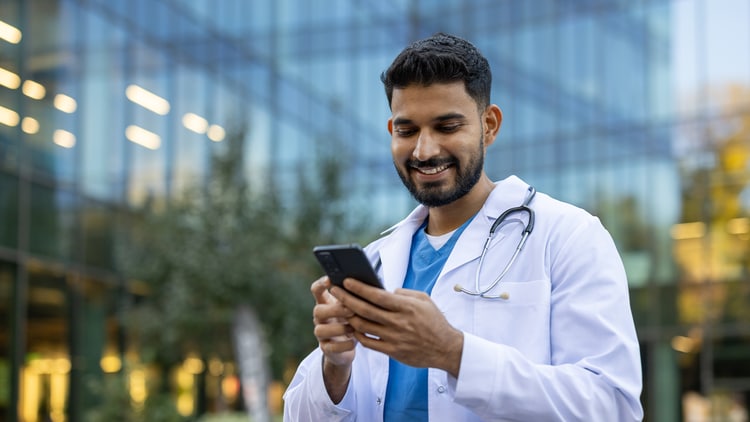 Innovationen im Gesundheitswesen: Mann in OP-Kleidung und Laborkittel lächelt vor einem hohen Gebäude in sein Telefon