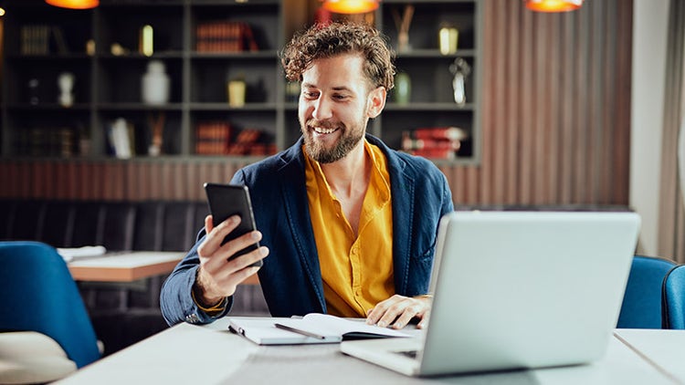 Geschäftsmann in Smart-Casual-Kleidung, der ein Smartphone benutzt, während er am Laptop arbeitet