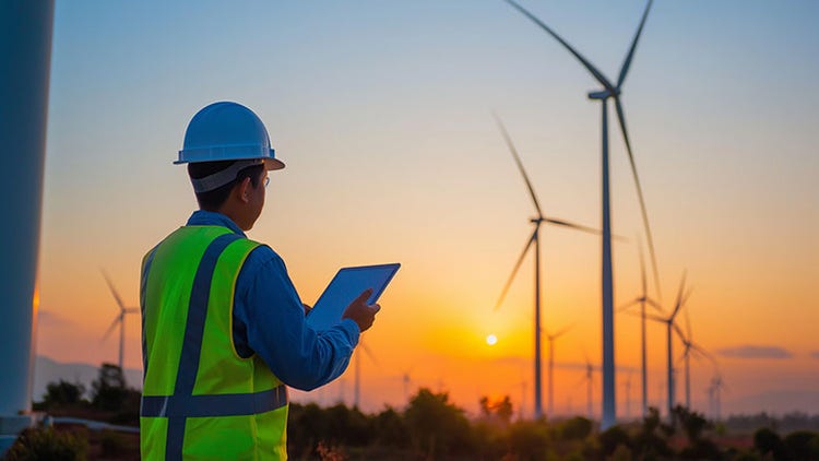 Homme portant un casque et un gilet de sécurité utilisant une tablette à l'extérieur face à des éoliennes