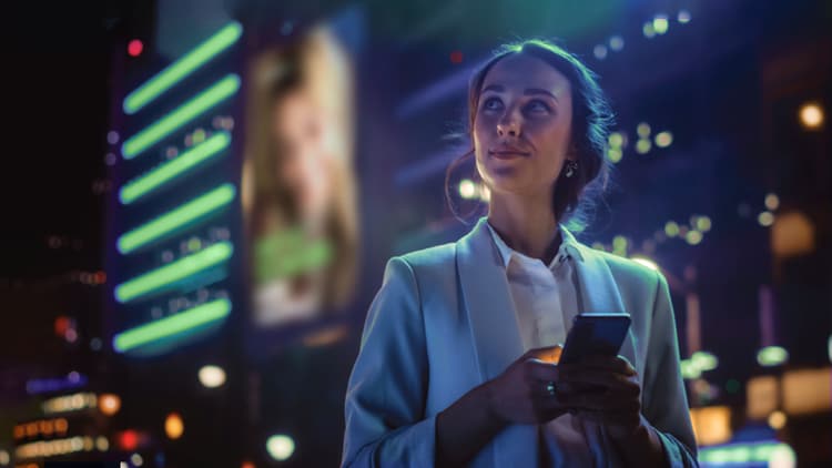 La révolution de l’IA : femme dehors avec un téléphone entre les mains, regardant au loin vers le haut et se tenant devant de grands bâtiments éclairés en pleine nuit