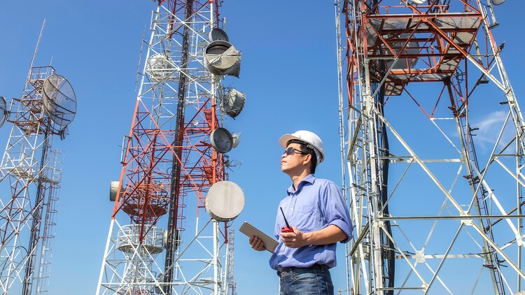 Telecommedewerker met een helm die een tablet vasthoudt en naar een aantal zendmasten kijkt