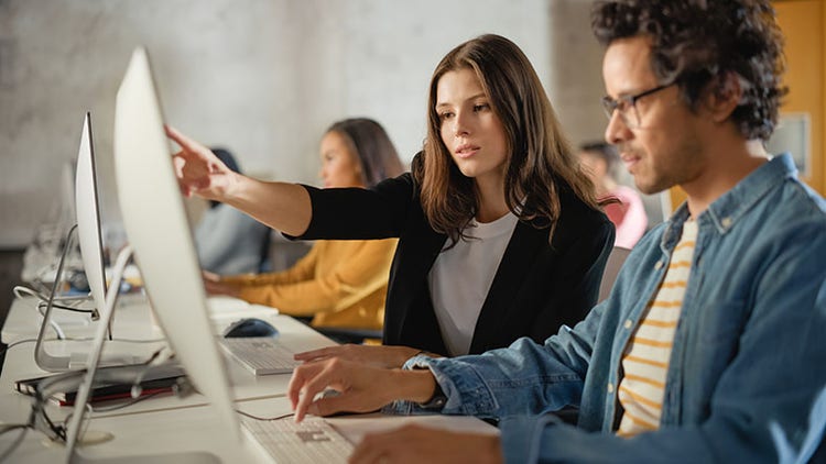 A woman pointing at a computer screen while assisting a man in an office setting