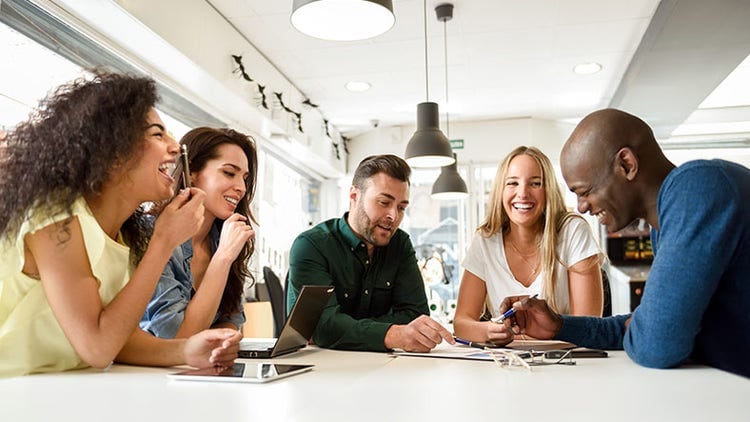 Five people gathered around a table, engaged in discussion with laptops and papers.