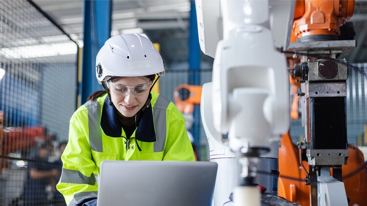 AI in manufacturing: worker in a safety helmet and jacket working on a laptop next to a robotic machine