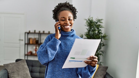 A woman smiling at home, holding a smartphone and reviewing paperwork
