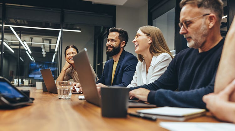 A group of professionals in a modern office, engaged in a meeting and smiling while using laptops