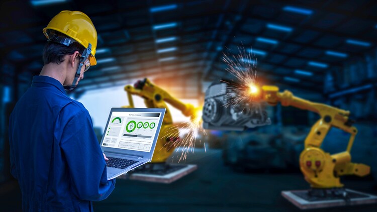 Helmeted man looking at a laptop in his hands in front of manufacturing machinery