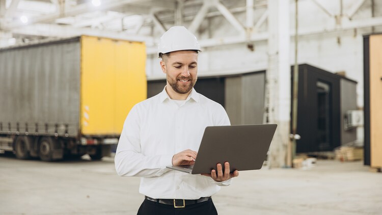 Agentic AI in manufacturing: man in a hard hat smiling at a laptop in his hands with an ariculated lorry in the background