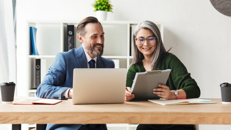Two smiling workers at a desk looking at an open laptop and paperwork in a bright office