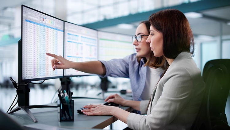 Two women analysing data on dual monitors, one pointing at a spreadsheet