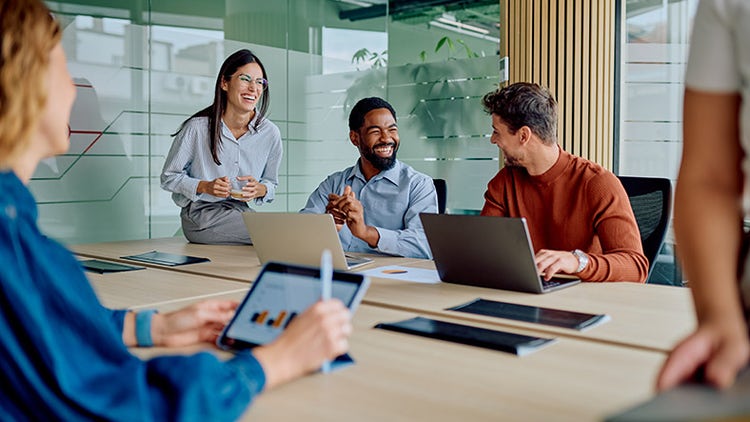 A diverse team of colleagues smiling and collaborating around a table of laptops in a bright, modern office meeting room.