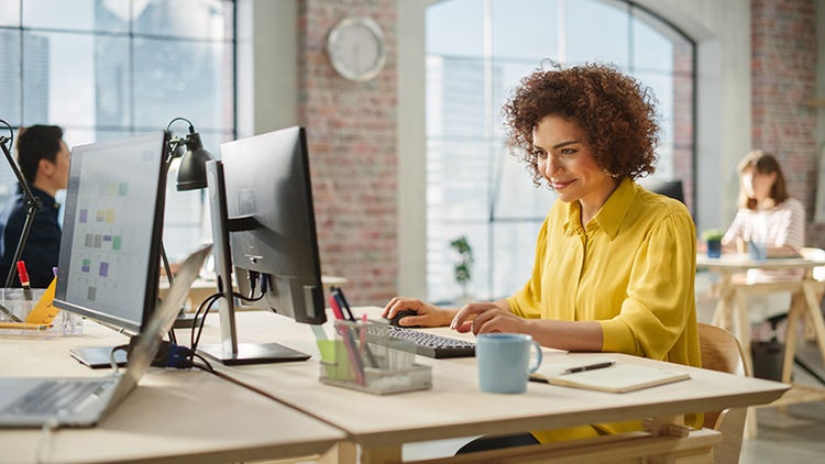 Woman in yellow shirt working at a desktop computer in a modern office with colleagues in the background
