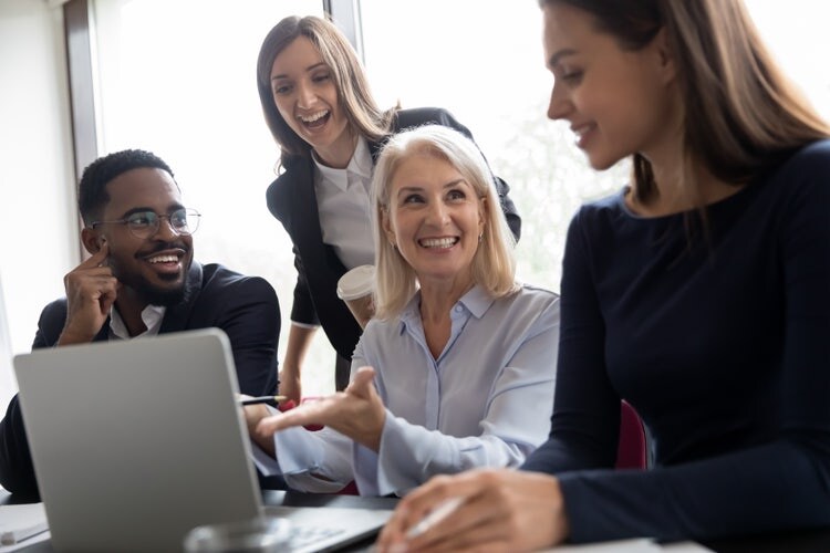 Four professionals gathered around a laptop, smiling and collaborating in an office